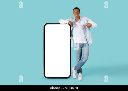 Smiling young african american guy in casual pointing finger at huge phone with blank screen, recommend new gadget Stock Photo