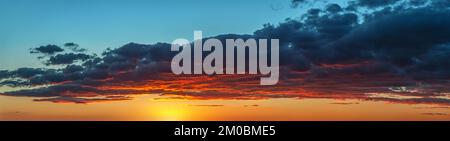 Setting sun painting red the bottom of large gray cloud. Dramatic sky at golden hour. Bright vivid sunset sky panorama. Picturesque wide skyscape. Stock Photo