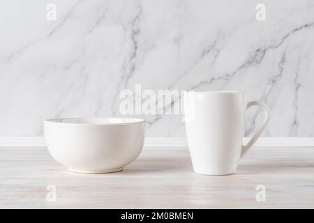 Bowl and mug on a table against marble wall. Blank ramekin and large cup mockup for food and drink design. Modern crockery and white tableware concept Stock Photo