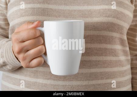 Woman hand holding white coffee mug. Female hand holds blank tea cup against striped wool sweater. Modern tableware, white crockery, hot beverage. Stock Photo