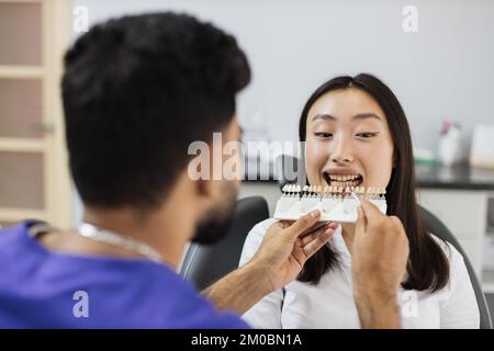 Dentist choosing color of filling from palette. Back view of male dentist checking and selecting color of young asian woman teeth indoors in modern hospital. Stock Photo