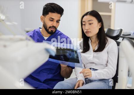 Medicine, dentistry and oral care concept. Male confident dentist showing x ray picture to attractive young patient, asian woman at modern light dental clinic. Stock Photo