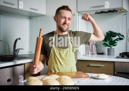 Portrait of caucasian baker man in green apron uniform smiling looking at camera showing biceps muscles. Handsome male baker at home sitting at the table with rolling pin. High quality photo Stock Photo