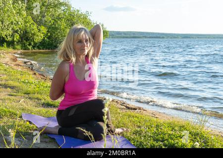 An elderly woman in sports clothes practicing yoga sitting cow pose and holding her hands folded together on the shore of the lake. Outdoor sports. Yo Stock Photo