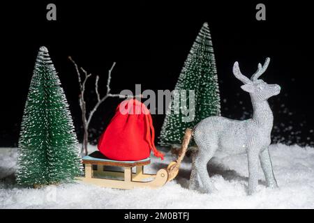 Creative Christmas card. A toy shining silver deer carrying a sleigh with a bag of gifts through the snowy forest among the Christmas trees at night. Stock Photo