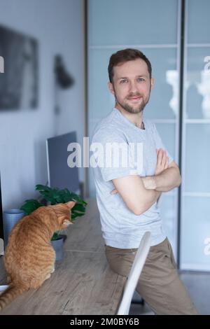 Portrait of cheerful bearded caucasian male freelance software developer at working place in home office near computer with tabby cat looking at camera. High quality vertical image Stock Photo