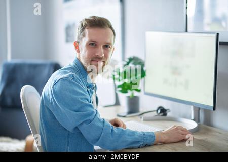 Young handsome IT support man in denim shirt . Attractive bearded caucasian male freelance software engineer sitting at working place in office hardly working using computer. High quality image Stock Photo