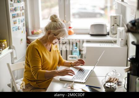 Mature woman working on laptop in kitchen Stock Photo