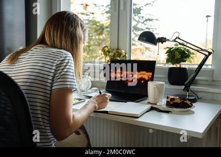 Freelancer working at home office. Back view of woman using laptop and writing in notepad sitting at table in home office Stock Photo