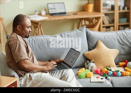 Side view portrait of single father working from home and using laptop on couch with toys, copy space Stock Photo