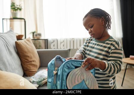 Portrait of cute black kid playing with toy bag in cozy home setting packing for nursery school, copy space Stock Photo