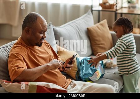 Side view portrait of father using smartphone on sofa at home with son playing in background, copy space Stock Photo