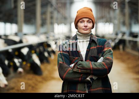Young successful female owner of cowfarm in workwear standing in front of camera with her arms crossed on chest against cowsheds Stock Photo