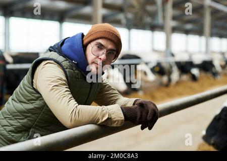 Young serious farmer in eyeglasses, beanie cap and workwear looking at camera while bending over cowshed against row of dairy cows Stock Photo
