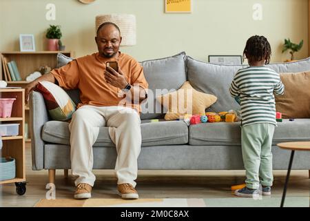 Full length portrait of father using smartphone with son playing on sofa at home, copy space Stock Photo