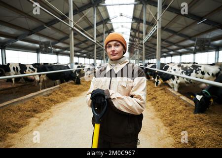 Young female owner of modern cowfarm in workwear holding woorktool while standing in front of camera in aisle between cowsheds Stock Photo