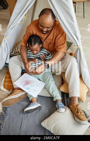 Vertical full length portrait of loving black father with child drawing together in play tent Stock Photo