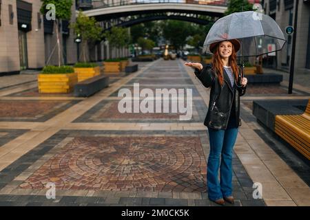 Full length portrait of positive young woman in elegant hat standing on European city street with transparent umbrella enjoying rainy weather outdoor Stock Photo