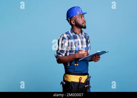 Portrait of construction contractor checking the task list on clipboard, looking carefully to make sure that all outstanding tasks have been completed. Construction worker against blue background. Stock Photo