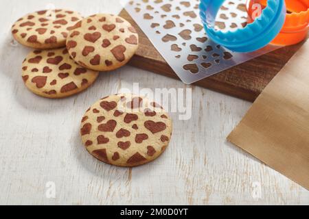Сookies with a pattern in the form of hearts, cookie cutters, stencil, cutting board and baking mat on a white wooden surface Stock Photo