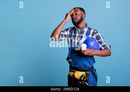 Exhausted construction employee drinking coffee during break. Overworked worker holding protective helmet wearing bibs while resting against blue background in studio shot. Stock Photo