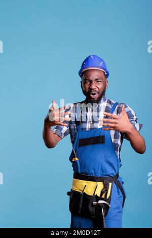 Construction employee dissatisfied with working conditions arguing with contractor. Upset african american worker, wearing work uniform and hard hat, studio shot against blue background. Stock Photo
