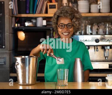 Portrait of smiling African American female bartender with afro hair looking at camera while serving cocktail Stock Photo