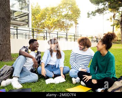 Group of diverse college student friends laughing and chatting sitting on the grass on campus Stock Photo