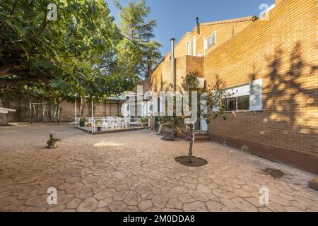 Facade of a detached house with a backyard with tiled floors, tree grates and fig trees and a dining room with a dining table and chairs Stock Photo
