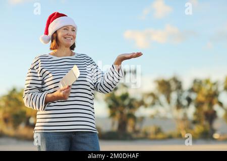 Adult smiling woman in Santa hat holds passport, ticket for trip and shows hand at copy space on palms trees background, New Year, Christmas winter ho Stock Photo