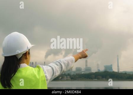 Female chief engineer wearing a green vest and helmet stands outside against the background of coal power plant station and steam in the morning mist. Stock Photo