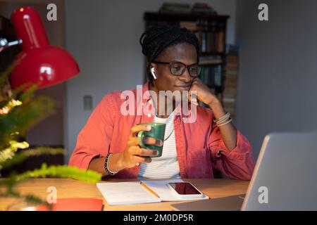 Smiling African student guy in wireless earbuds drinking tea while watching online lectures at home Stock Photo