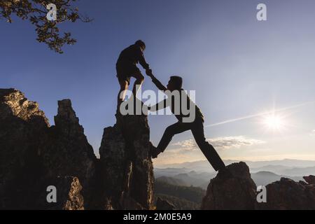 Silhouette of  two people climbing on mountain cliff and one of them giving helping hand. People helping and, team work concept. Stock Photo