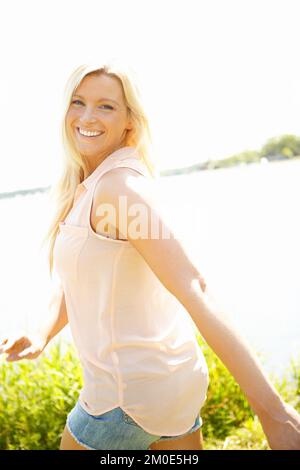 I love the outdoors. Portrait of a smiling young blond woman walking near a lake. Stock Photo