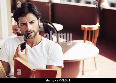 Hes a real man about town. Young handsome man smoking a pipe and reading a book at a cafe. Stock Photo