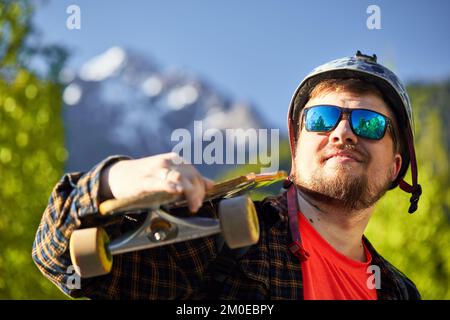 Portrait of serious bearded man Skater with sunglasses and helmet holding his longboard at mountain background Stock Photo
