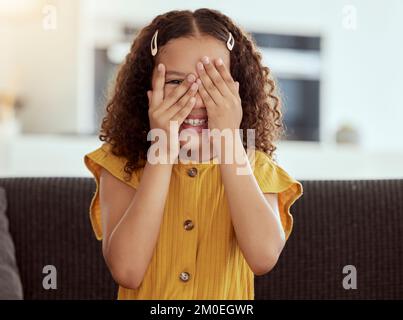 Adorable little mixed race child covering face with hands and peeking. One small cute hispanic girl sitting alone on living room sofa and playing hide Stock Photo