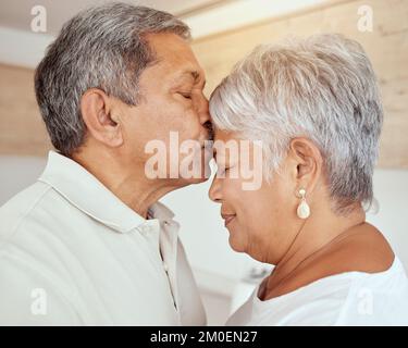 Closeup of mixed race senior man kissing wifes forehead in the morning at home. Affectionate elderly husband and wife standing and bonding in kitchen Stock Photo