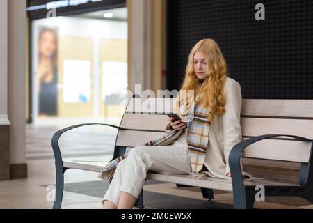 Young beautiful woman in light coat sitting on the bench inside a shoping mall and texting with a smartphone. Exhausted young woman tired from shoppin Stock Photo