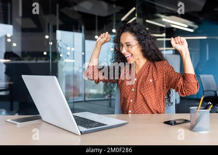 Hispanic business woman celebrating victory success, employee with curly hair inside office reading good news, using laptop at work inside office holding hand up and happy triumph gesture. Stock Photo