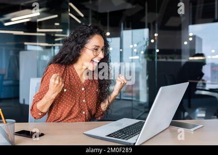 Hispanic business woman celebrating victory success, employee with curly hair inside office reading good news, using laptop at work inside office holding hand up and happy triumph gesture. Stock Photo