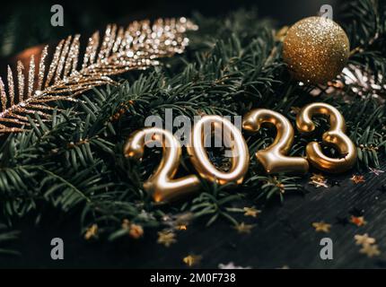 Close-up still life photo of golden numbers and decorations for new year 2023. Gold bauble and decorations on dark green pine branch. Stock Photo