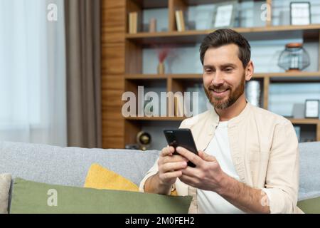 A mature man with a beard in a shirt at home is sitting on the sofa in the living room, using the phone, typing messages and browsing the Internet online. Stock Photo