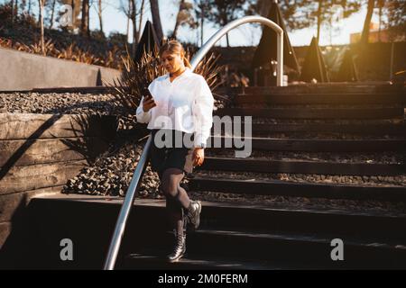 A plus-sized woman dressed in an oversized white shirt, black button-down skirt, tights, and black military boots; Walks down the stairs of a garden h Stock Photo