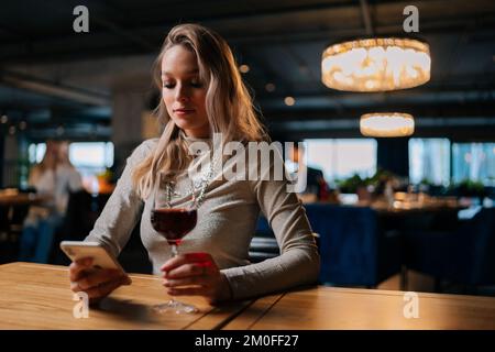 Portrait of serious young woman using smartphone, typing online message sitting at table holding in hand glass of red wine at restaurant with dark Stock Photo
