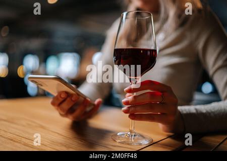 Close-up cropped shot of unrecognizable young woman using smartphone, typing online message sitting at table holding in hand glass of red wine at Stock Photo