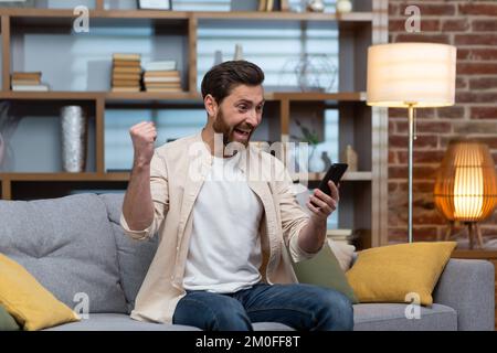 Man at home on sofa using smartphone, celebrating victory in living room reading happy news notifications from phone online holding hand up gesture of triumph and success sitting on sofa. Stock Photo