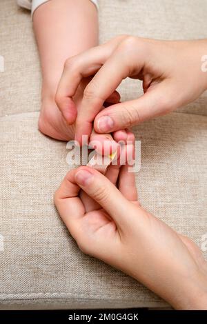 Mother woman sticks a medical adhesive plaster on the toddler baby leg. Mom s hand with sticky wound protection tape and child s foot. Kid aged one ye Stock Photo