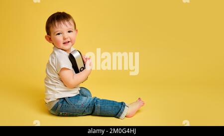 Toddler baby plays with a wireless music speaker on a studio yellow background. Happy child in a blue jeans listens to music in an audio speaker, copy Stock Photo