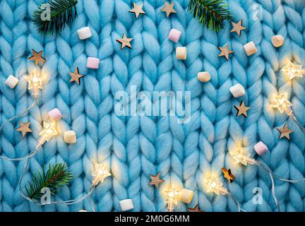 Flat lay view of light blue soft knitted pattern wool background with star shape string lights, spruce tree branch and mini marshmallows, Christmas. Stock Photo